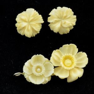 Puede incluir: Un conjunto de tres broches de flores talladas en marfil. Dos broches más pequeños son diseños de flores simples, mientras que el broche más grande presenta una flor más detallada con un círculo central.