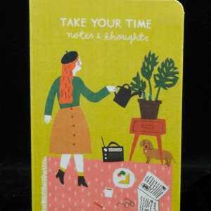 May include: A yellow notebook with the text "TAKE YOUR TIME notes & thoughts" on the cover. The cover features an illustration of a woman watering a plant, a dog, and other objects on a pink and white patterned rug.