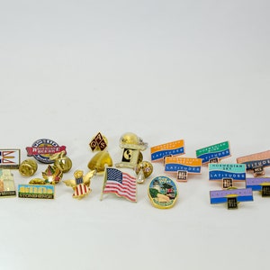May include: A collection of 20 enamel pins featuring various designs, including flags, buildings, and logos. The pins are gold-toned with colorful enamel accents. Some of the pins feature text, such as "Adventure Ocean", "Stonehenge", and "Norwegian Sky".