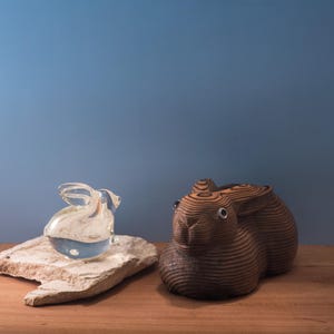 May include: A clear glass rabbit sculpture with a curved handle sits on a beige stone. Next to it is a wooden rabbit figurine with a layered design. Both are on a wooden surface against a blue background.