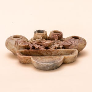 May include: A carved stone sculpture with a light brown and reddish-brown color palette. The sculpture features three vase-like structures and a central floral design. The base has a curved, tiered design. The piece is likely a decorative item.