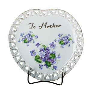 May include: White porcelain plate with a gold trim and a scalloped edge. The plate features a floral design with purple flowers and green leaves. The words "To Mother" are written in gold script on the plate.