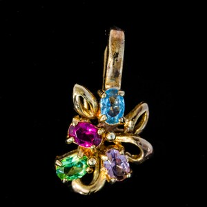 Peut inclure: Pendentif doré avec un motif floral orné de cinq pierres précieuses ovales de couleur bleue, fuchsia, verte et violette. Le pendentif est doté d'une boucle pour être suspendu, sur fond noir.