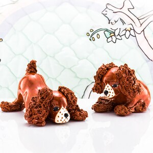May include: Two brown ceramic dog figurines with curly fur and white and black facial markings. One dog is lying down, and the other is sitting. The background features a light green and white patterned design.