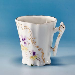 May include: White porcelain teacup with a scalloped rim and handle. The cup is decorated with purple and yellow floral designs and gold accents. The handle has a decorative shape.