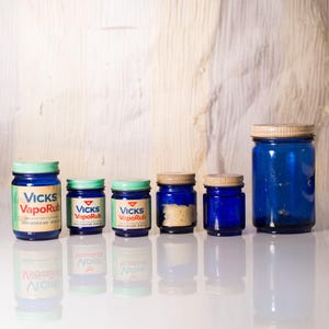 Könnte beinhalten: Fünf blaue Glasgefäße mit grünen Deckeln. Das größte Gefäß ist leer, während die vier kleineren Gefäße Etiketten mit der Aufschrift "Vicks VapoRub" tragen.