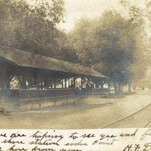 May include: Sepia-toned vintage photograph of a lakeside structure with a covered roof, likely a train station or pavilion. A road and train tracks run alongside, with trees lining the path. Handwritten text is visible at the bottom.