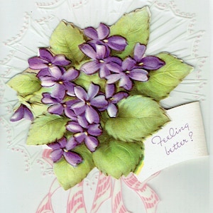 May include: A vintage greeting card with a bouquet of purple flowers and green leaves. The card has a white background with a pink and white embossed design. The text on the card reads "Feeling better?"
