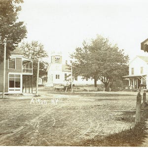 May include: Sepia-toned photograph of a small town square with buildings and trees. A two-storey building with a porch and a church with a steeple are visible. The text "Alton NY" is written on the ground.