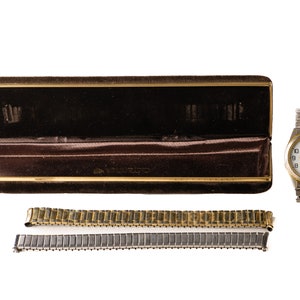 May include: A gold-tone watch with a white face and a date window. The watch comes with a brown velvet box and two metal watch bands, one gold-tone and one silver-tone.