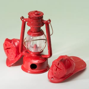 May include: A red toy lantern with a clear glass globe and a handle, accompanied by two red toy firefighter helmets. The helmets have the text "18" and "Fire Company" on them. The lantern is a classic design.
