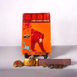 Puede incluir: Libro vintage "Bear Cub Scout Book" con una portada roja y naranja con una ilustración de oso. Incluye una hebilla de cinturón, cinturón y varios alfileres. El libro y los accesorios se exhiben sobre una superficie blanca.