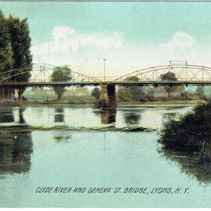 May include: A vintage postcard showing the Clyde River and Geneva St. Bridge in Lyons, N.Y. The bridge is a steel arch structure over a calm river, with trees and foliage on the banks. The sky is a pale blue.