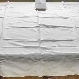 Könnte beinhalten: Weiße Tischdecke mit einem zarten Stickmuster an den Rändern. Die Tischdecke liegt auf einem Holztisch.