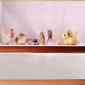 May include: A display shelf featuring a collection of bird figurines. The figurines include a white chicken with a red comb, a brown bird, a small bird with a black base, a ceramic owl, and a yellow owl with wings. The background is a patterned fabric.