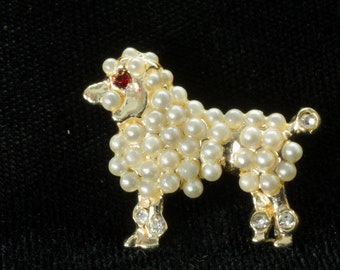 May include: A gold-toned brooch shaped like a poodle, covered in white faux pearls with a single red rhinestone eye.