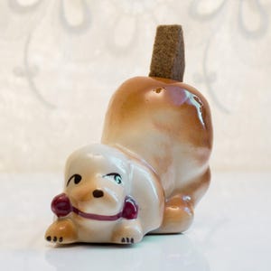 May include: A ceramic dog figurine in a playful pose, featuring a light beige body with brown accents. The dog has a red collar and a brown, rectangular stopper on its back. The figurine is on a white surface.