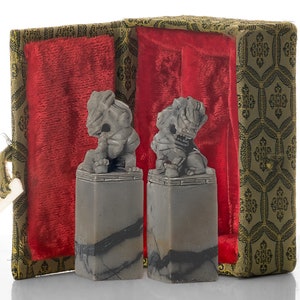 May include: Two gray stone carvings of lions, each sitting on a square base, are displayed in a red velvet lined box with a gold patterned exterior.