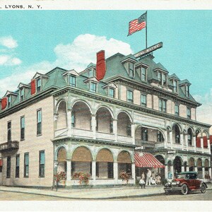 May include: Vintage postcard of the Hotel Wayne in Lyons, N.Y. The multi-story building features a red, white, and blue American flag, a red awning, and a classic car parked out front. The sky is blue with a few clouds.