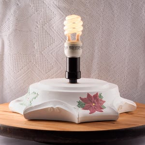 Könnte beinhalten: Ein weißer Keramik-Lampensockel mit schwarzem Lampenhalter und einer Spirallampe. Der Sockel hat eine sternartige Form mit einem roten Weihnachtsstern und grünen Blättern. Die Lampe steht auf einer Holzoberfläche.