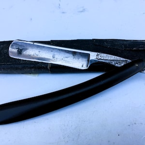May include: A straight razor with a black handle and a silver-coloured metal blade. The blade features an etched inscription that reads Made From Kayser Ellison Best Sheffield Silver Steel. The razor is accompanied by a black, rectangular protective case.