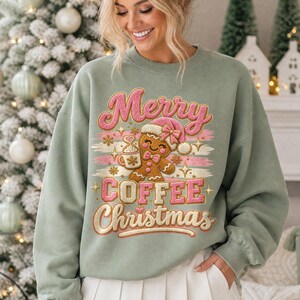 May include: A sage green crewneck sweatshirt featuring a graphic of a gingerbread person holding a coffee mug, surrounded by pink and gold text that reads Merry COFFEE Christmas, with decorative stars and horizontal brushstroke accents.