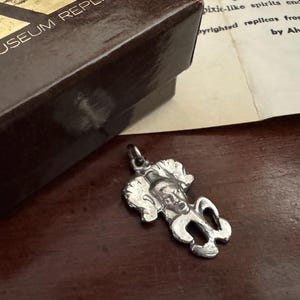 Pode incluir: Um pingente prateado em forma de gremlin, ao lado de uma caixa marrom com o texto "ALVA MUSEUM REPLICAS" e um papel com texto sobre o pingente. O pingente tem um rosto detalhado e cabelo encaracolado.