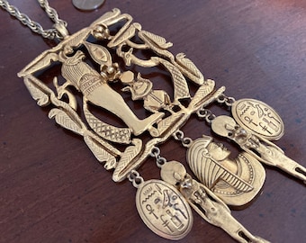 Puede incluir: Un collar con un colgante cuadrado dorado de temática egipcia, que presenta un recorte detallado de una figura de pie con jeroglíficos. El colgante está unido a una cadena con varios colgantes más pequeños, cada uno con un diseño jeroglífico diferente.