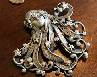 May include: A silver-toned brooch featuring a woman's face with flowing hair adorned with stars and rhinestones. The design is intricate and detailed, with a whimsical and ethereal feel.