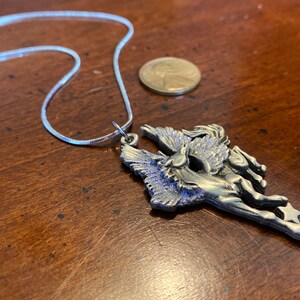May include: A silver-toned Pegasus pendant necklace. The pendant features a detailed winged horse design with blue accents. The necklace includes a thin, silver chain. A penny is next to the necklace.
