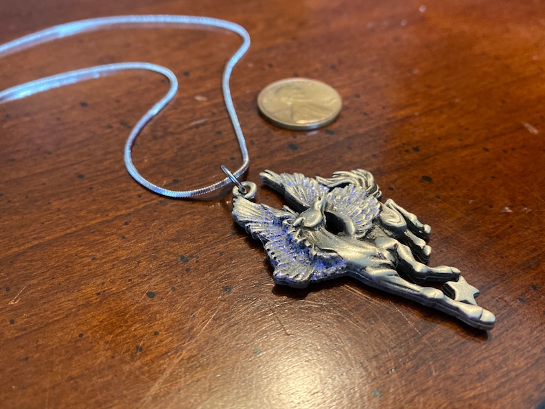 May include: A silver-toned Pegasus pendant necklace. The pendant features a detailed winged horse design with blue accents. The necklace includes a thin, silver chain. A penny is next to the necklace.