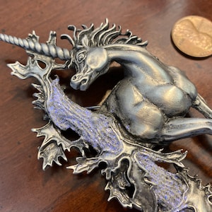 May include: A silver unicorn brooch with a detailed design. The unicorn is standing on a leafy branch with a sparkly blue finish.