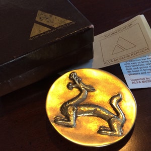 May include: A gold-colored metal dragon sculpture on a round, gold-colored metal base. The sculpture is a replica of an original piece and is part of a collection of museum replicas. The text on the card reads "ALVA MUSEUM REPLICAS".