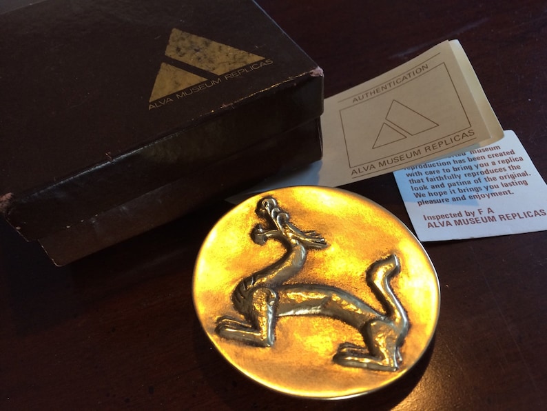 May include: A gold-colored metal dragon sculpture on a round, gold-colored metal base. The sculpture is a replica of an original piece and is part of a collection of museum replicas. The text on the card reads "ALVA MUSEUM REPLICAS".