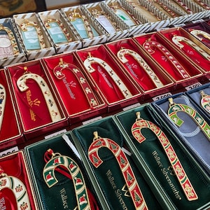 May include: A collection of vintage candy cane Christmas ornaments in red, green, and white boxes. The ornaments are made of metal and feature a gold finish. The boxes are decorated with the Wallace Silversmiths logo and the year the ornament was made.