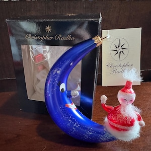 Puede incluir: Adorno navideño de luna creciente azul con una cara sonriente y detalles de purpurina. Una pequeña figura de Papá Noel está posada en la luna. El adorno está junto a una caja negra y una etiqueta con el nombre "Christopher Radko".