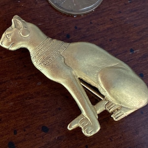 May include: A gold-colored metal brooch in the shape of a seated cat. The cat has a detailed design with a collar and a decorative pattern on its back.