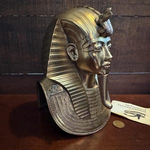 May include: Gold-colored bust of King Tutankhamun, wearing a traditional pharaoh's headdress with a cobra on the forehead. The bust is on a wooden surface with a small paper tag that reads "The Treasures of Tutankhamun".