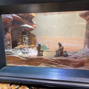 May include: A framed diorama depicting a desert scene with two figures, a ladder, and a small building. The figures are made of wood and are painted in brown and black. The diorama is set against a backdrop of a desert landscape with a light brown and pink sky.