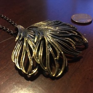 May include: A gold-toned necklace with a large, intricate pendant shaped like a stylized leaf. The pendant is made of multiple layers of metal, creating a textured and dimensional look.