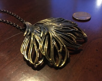 May include: A gold-toned necklace with a large, intricate pendant shaped like a stylized leaf. The pendant is made of multiple layers of metal, creating a textured and dimensional look.