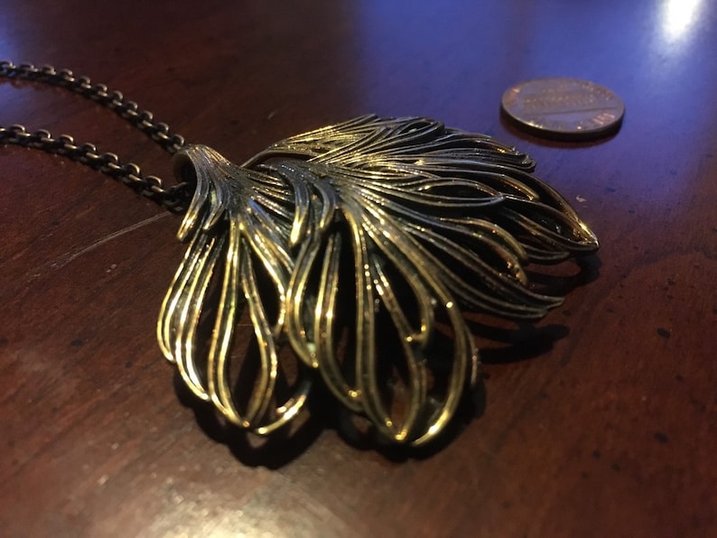 May include: A gold-toned necklace with a large, intricate pendant shaped like a stylized leaf. The pendant is made of multiple layers of metal, creating a textured and dimensional look.