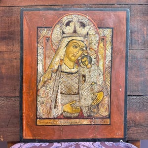 May include: Antique religious icon featuring a depiction of the Virgin Mary holding the Christ Child. The artwork is painted in warm tones of gold, red, and brown, with intricate details and text at the bottom. The icon is framed in a reddish-brown border.
