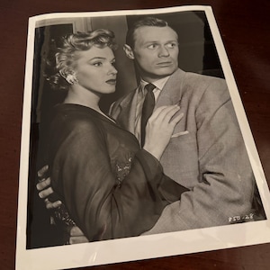 May include: Black and white photograph of a man and woman standing close together. The woman is wearing a dark dress and the man is wearing a suit jacket and tie. The man has his arm around the woman's waist.