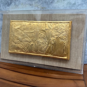May include: A gold-coloured rectangular relief, depicting figures and hieroglyphs, is encased in a clear acrylic box. The relief is mounted on a beige fabric background. The artwork is displayed on a wooden surface.