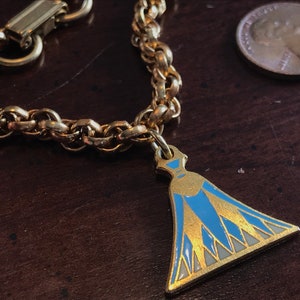 May include: A gold chain necklace with a blue and gold pendant shaped like a dress. The pendant has a white and blue geometric design.