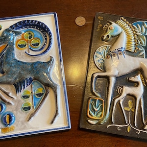 May include: Two ceramic wall plaques with a blue and white stylized goat on one and a white horse with a foal on the other. Both plaques have a dark brown background with stylized floral designs.