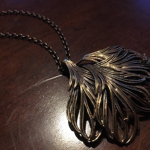 May include: A gold-toned necklace with a large, intricate pendant shaped like a stylized feather. The pendant is made of multiple layers of metal that create a delicate, airy design.
