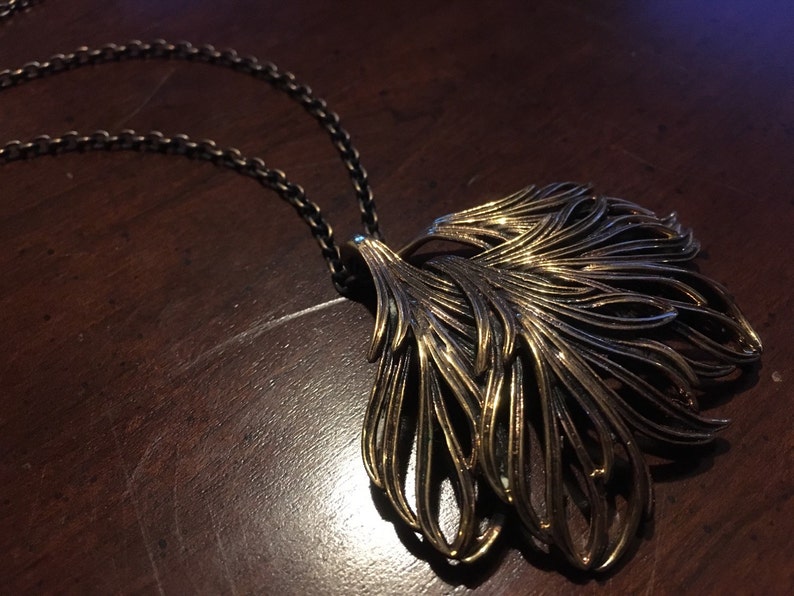 May include: A gold-toned necklace with a large, intricate pendant shaped like a stylized feather. The pendant is made of multiple layers of metal that create a delicate, airy design.