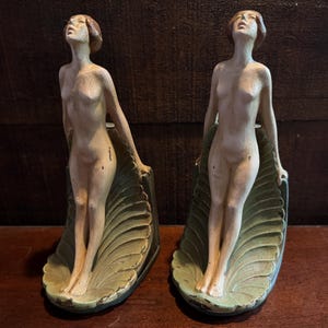May include: Two antique statues of nude figures, each standing on a stylized leaf base. The figures have their heads tilted upwards. The statues are painted in muted tones of beige, green, and brown, with visible signs of age and wear.